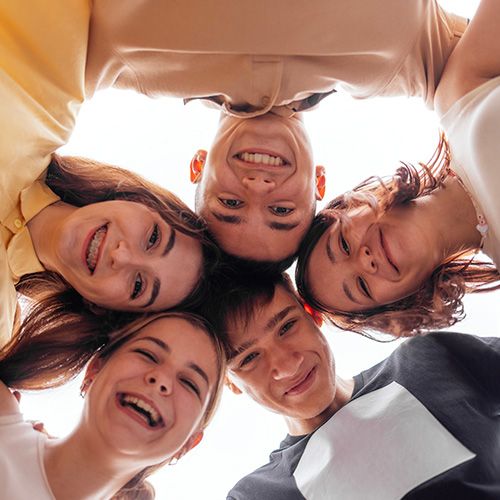 Una fotografía tomada desde un ángulo bajo de un grupo de cinco jóvenes sonriendo y mirando hacia la cámara. Están en un círculo muy unido, y se pueden ver sus rostros alegres.