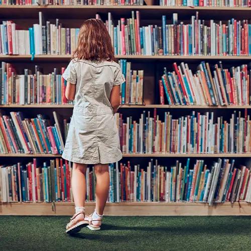 Una niña de pie de espaldas, mirando una estantería alta y llena de libros en una biblioteca.