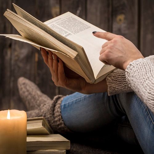 Una persona sentada contra una pared de madera, con las piernas extendidas y cubiertas con calcetines de punto y jeans, leyendo un libro abierto. Una vela encendida y una pila de libros están a la izquierda.