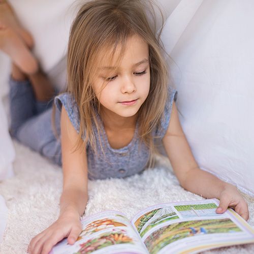 Una niña pequeña con cabello rubio claro y un atuendo casual de dos piezas, recostada boca abajo sobre una alfombra de piel sintética blanca, leyendo un libro abierto.