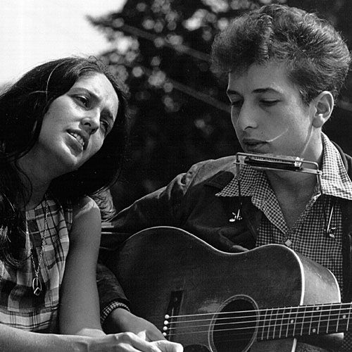 Una fotografía en blanco y negro de una mujer con cabello oscuro y un vestido a cuadros mirando hacia arriba y a la izquierda, mientras un joven con cabello rizado y una camisa a cuadros toca una guitarra acústica y una armónica simultáneamente. Parecen estar cantando juntos.